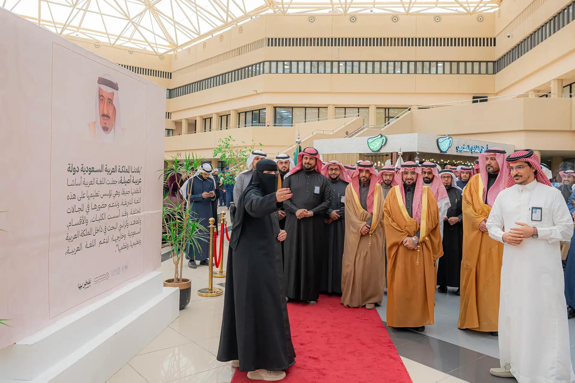 تجمّع عدد من الرجال بزيهم السعودي الأصلي، الحضور الشماغ أو الغترة السعودية، داخل قاعة عالمية بالقرب من سجادة حمراء في إطار نظمها الفعالة جامعة القصيم احتفاء باليوم للغة العربية. واستمع إلى كلمة ألقاها أحد الوجهين، بينما يأتي على الجانب المعماري لوحة عرض كبيرة تتضمن نصوصًا باللغة العربية وصورة شخصية، وذلك من خلال وسط المدير المعماري.