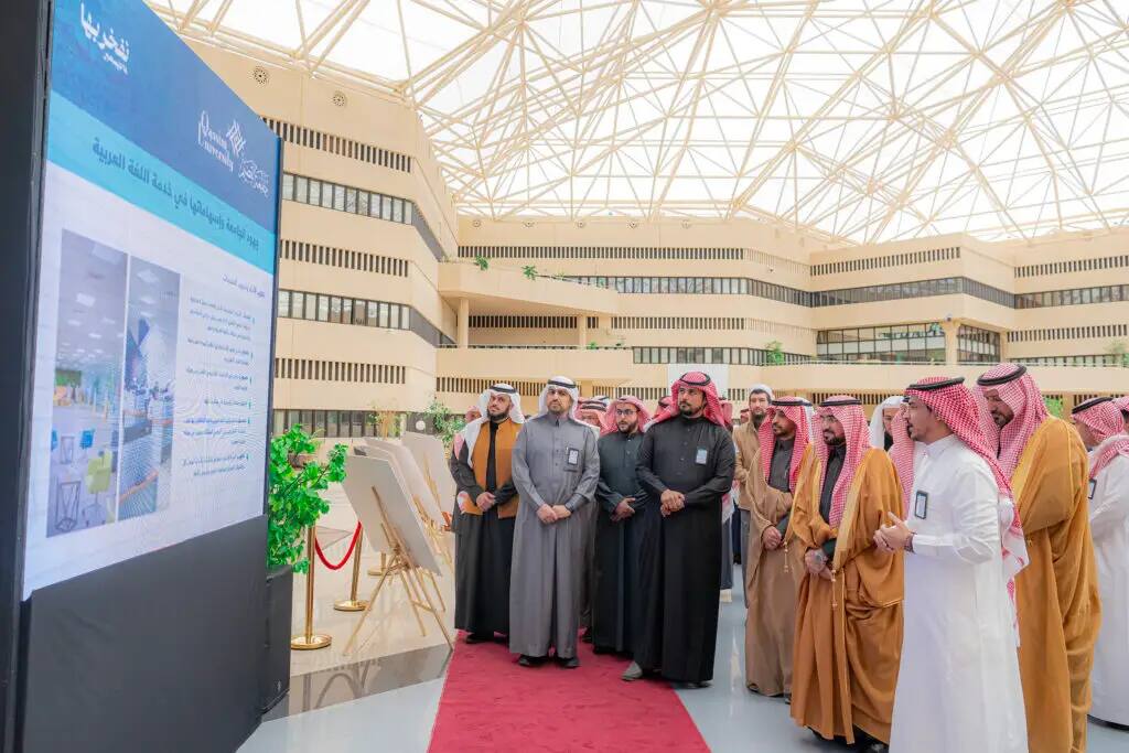 مجموعة رجال مرتدين الشماغ السعودي والغترة السعودية داخل قاعة مفروشة بالسجاد الأحمر، يتابعون باهتمام شاشة عرض رقمية كبيرة وتحديد اللغة العربية وتعرض صوراً متنوعة، وذلك وذلك في أحد أجنحة الجامعة الحديثة ذات القبة الزجاجية. يأتي ذلك احتفاءً باليوم العالمي للغة العربية في جامعة القصيم.