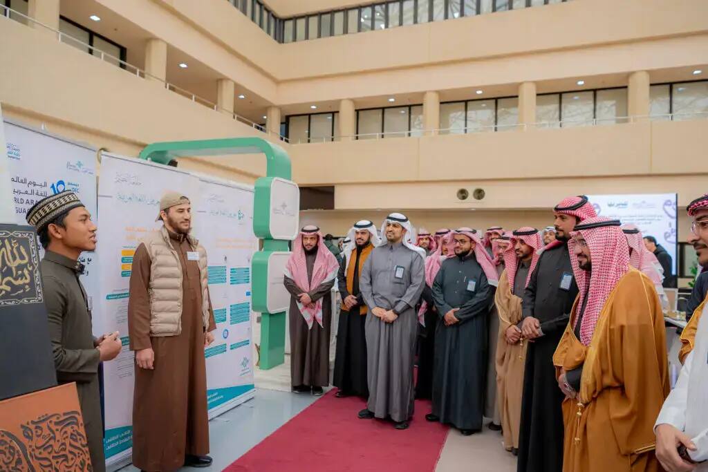 مجموعة الاتصال من الرجال الشماغ السعودي أو الغترة السعودية في قاعة الملابس الداخلية الشاملة وحديثة في القصيم، مصغين إلى المتحدث الشهير ينضم إليهم معلومات حول اللغة العربية معروضة على لوحات عرض، بينما يجتمع بعض الحضور فوق بساط أحمر.