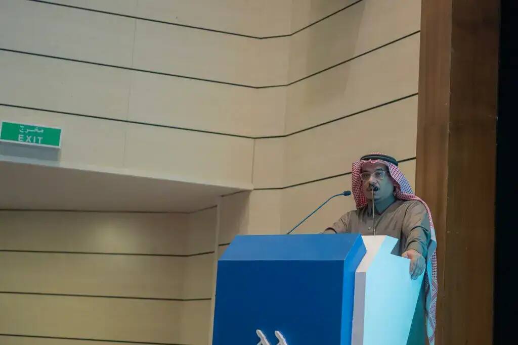 رجل يتحدث مرتدياً الثوب والشماغ السعودي في منصة الخطابة في قاعة الحديث في القصيم، وخلفه أخضر لمخرج الطوارئ، وذلك عبر فعالية اليوم العالمي للغة العربية.