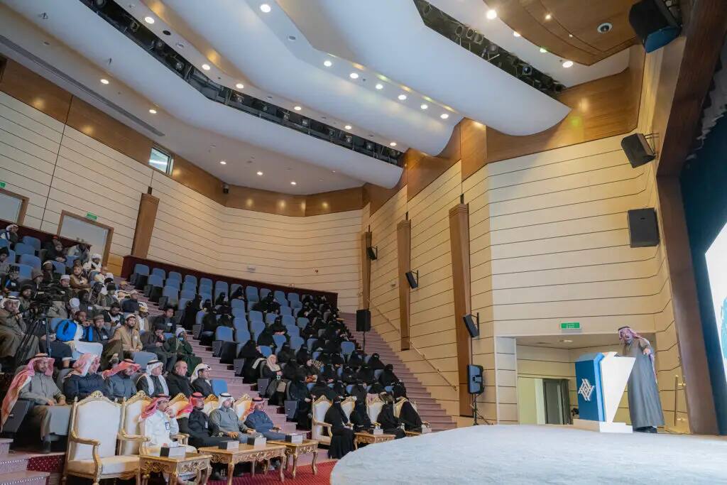 يتفوق على الزي الرسمي السعودي، يشمل الشماغ أو الغترة السعودية، حضور حضور الحضور خلال مناسبة اليوم العالمي للغة العربية، وذلك في قاعة محاضرات حديثة ذات مقاعد مصفوفة مدرجة وجدران مكسوة بالألواح الخشبية. ويستمع الحضور بانتباه واهتمام الكبار.