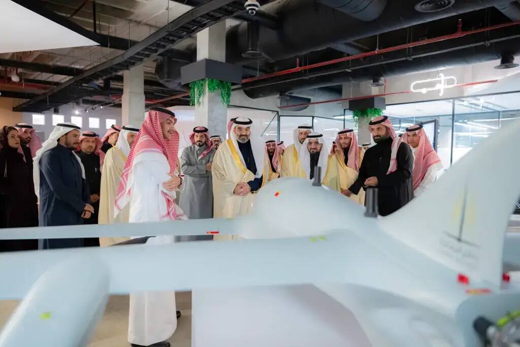 A group of men in authentic Saudi dress, including the Minister of Communications, gather around a large black drone on display inside a modern hall at Qassim University, where they learn about its features and research its details. Given the presence of those wearing the Saudi shemagh or Saudi ghutra in keeping with the use of patriotism, this certainly implies a technological advancement in the university's gambling.