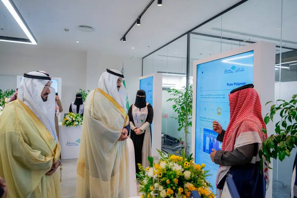 A group of men wearing the Saudi Shemagh or Ghutra, along with a number of women wearing abayas, around an informational display stand at the event as well as a comprehensive search. The modern venue, with digital lighting and flowers, reflects the modern feel of the event at Qassim University.