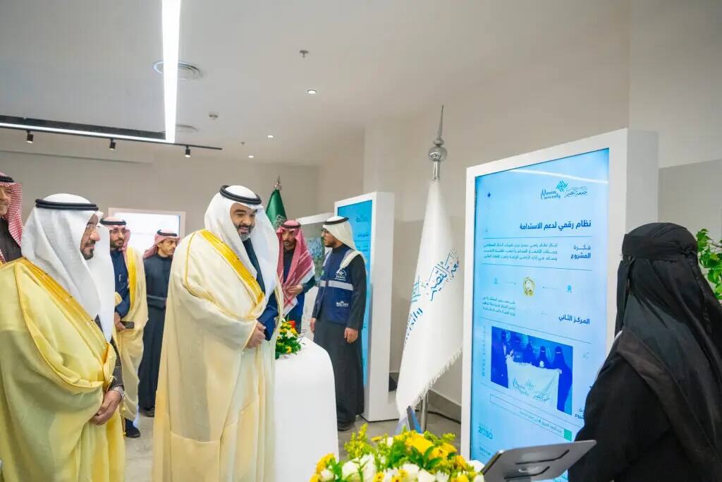 The image shows a number of men in traditional Saudi dress, including a shemagh or Saudi ghutra, as well as a woman dressed in white, looking at a digital screen during an event to search for verses. The background includes Qassim University, the Saudi flag and a number of informational posters.