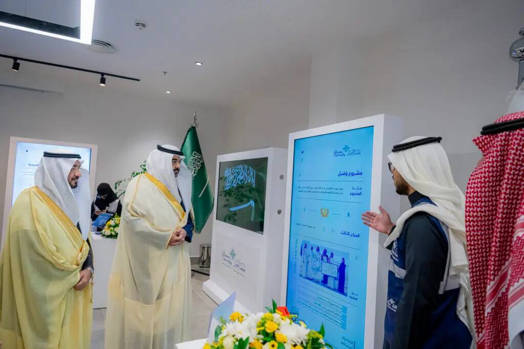 A number of attendees, dressed in Saudi Arabian shemagh and traditional attire, led by the Minister of Communications, watch a digital presentation in a modern hall topped by the flag of the Kingdom of Saudi Arabia. The neighboring format is one that embodies the interest in comprehensive research at Qassim University.