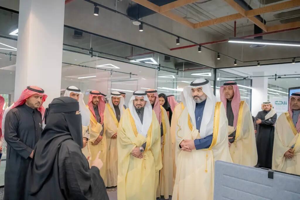A group of men dressed in formal attire with a Saudi shemagh or Saudi ghutra, listen intently to a woman dressed in black and a niqab, inside a modern, tiled office at Qassim University, characterized by bright lighting and geometric designs.