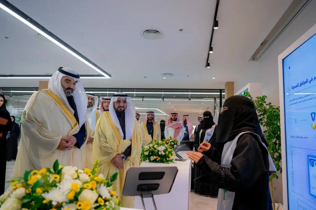 A group of men dressed in Saudi Arabian shemagh or ghitra with the traditional thobe, are listening intently to a woman presenting at a technology platform, wearing a black abaya and niqab, as she demonstrates a live search. Consider the modern setting of flowers and a large digital screen, reflecting the sophisticated nature of the event at Qassim University.