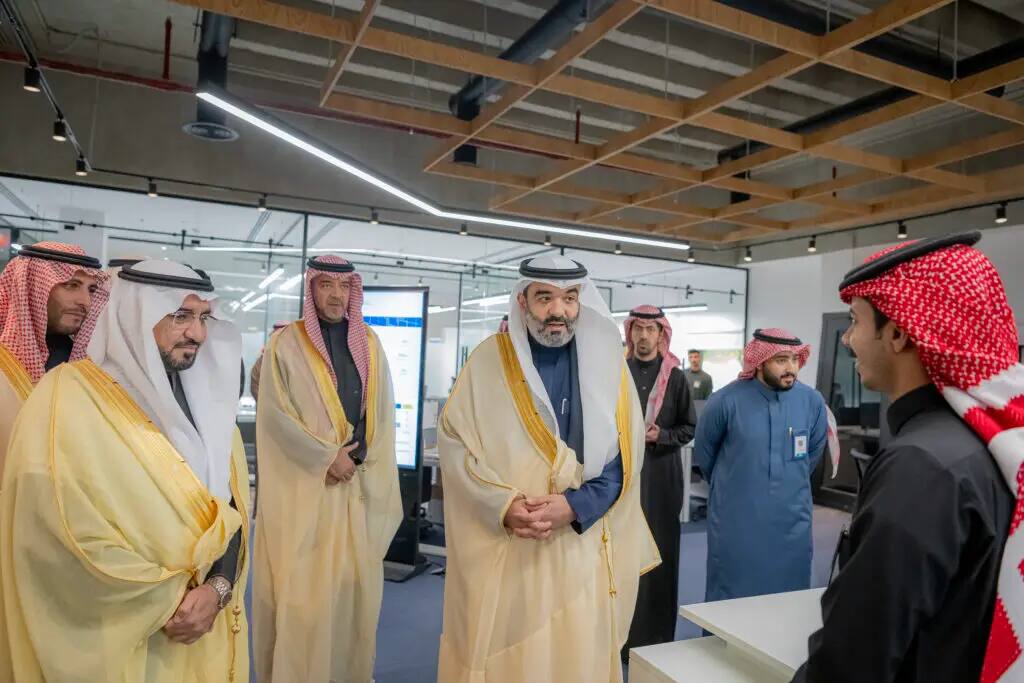 Of course, here is an official and appropriately paraphrased version based on your guidance: "A group of men in traditional Saudi dress, including a representative from Qassim University wearing a Saudi Shemagh or Ghutrah, are engaged in a discussion in a modern office environment. One person stands opposite the group at the desk, listening intently. The space, which features glass walls and digital screens, reflects an atmosphere dedicated to research and innovation.