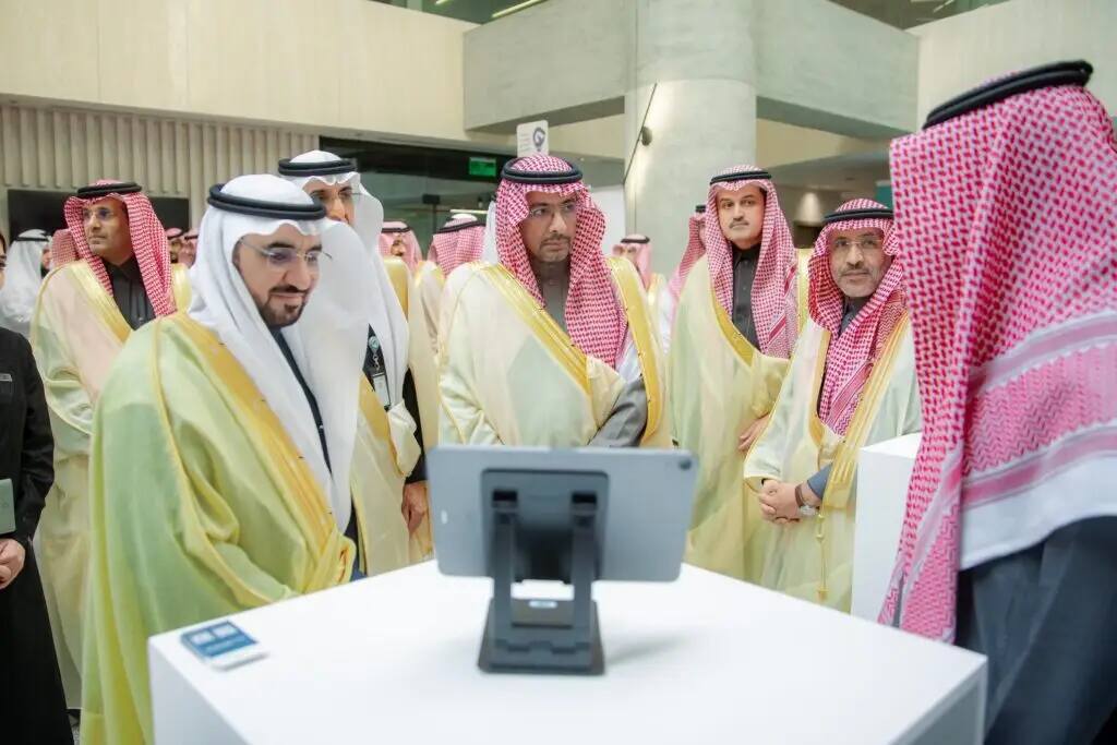 The caller is a group of men dressed in traditional Saudi attire, including white robes and a shemagh or Saudi ghutra, around a tablet screen during an industry event, watching the screen content intently and attentively.