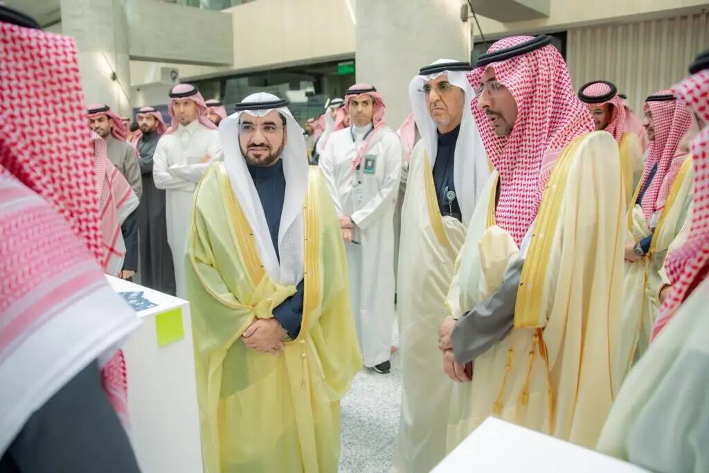 A number of men in traditional Saudi attire are gathered in one of the interior halls, dressed in robes and a Saudi jacket or shemagh. They gather around white tables and chat on the topic of industry and mining, while a moderator follows the discussion in the background.