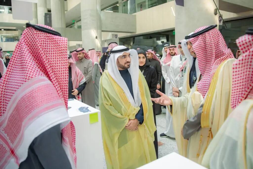 A group of men in authentic Saudi Arabian dress, including the thobe and shemagh or ghutra, gather and discuss issues related to industry and mineral wealth as part of an event or exhibition organized by Qassim University for children.