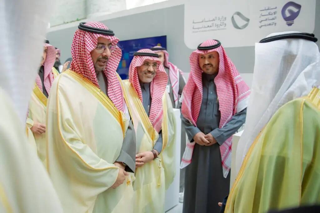 A group of men dressed in authentic Saudi dress, including red Saudi Arabian robes and shemaghs inside, chatting at an indoor event for makers and innovators, where I noticed a note in Arabic in the background.