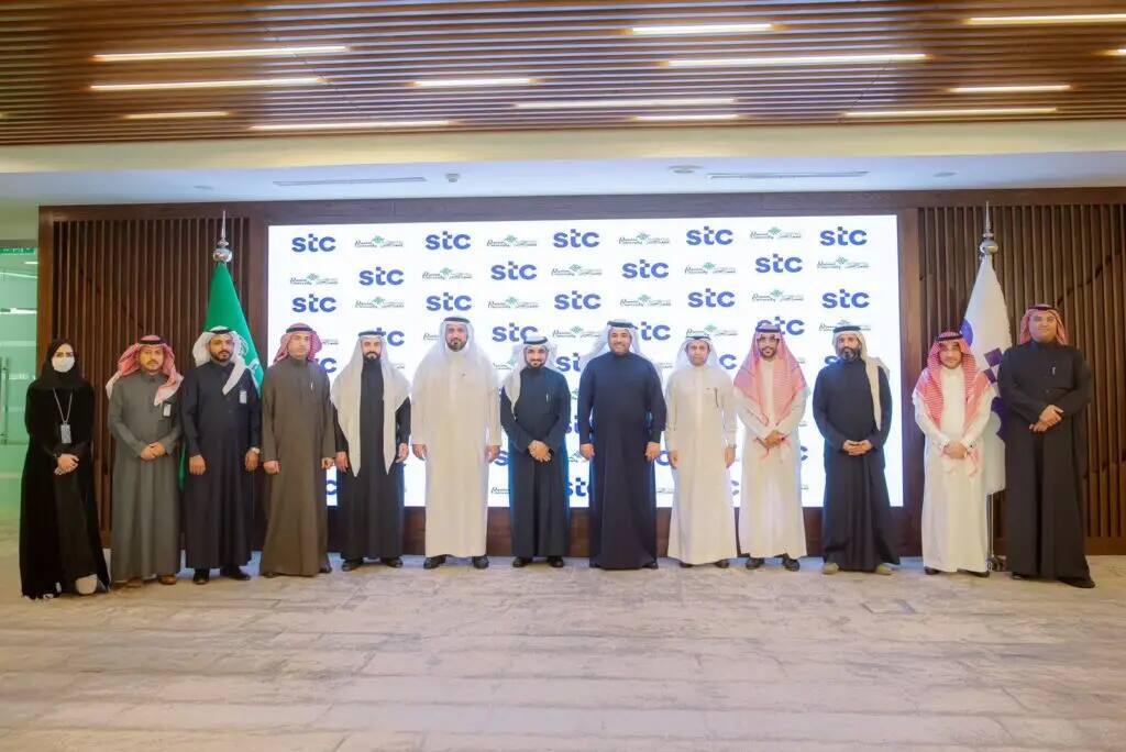 Six people, including men in the formal Saudi presence of the Saudi Shemagh or Saudi Ghutra, and women in formal attire, line up in front of a backdrop of STC Group logos and flags. The initiative is a memorandum signing ceremony between STC Group and Qassim University.