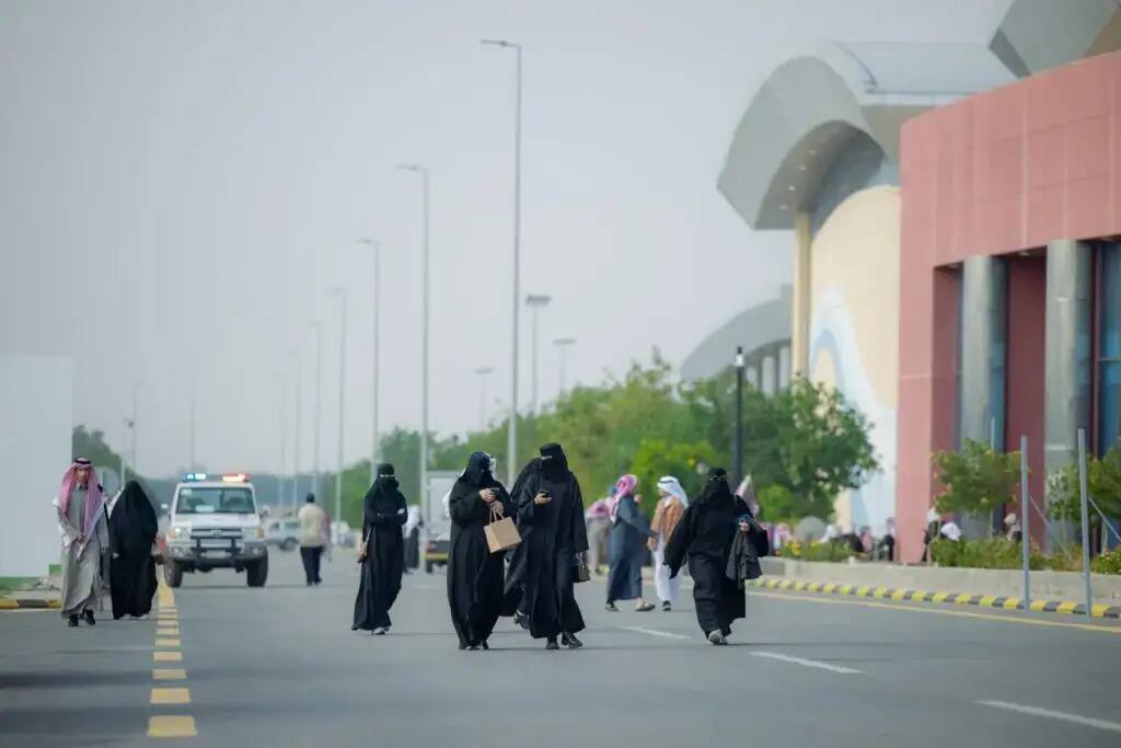 A group of people, including a number of women wearing black headscarves and niqabs, walk along the side of the road next to Qassim Modern University. A patrol car and a number of pedestrians are identified in the background. Some of the men can also be seen wearing the Saudi shemagh or ghutra.