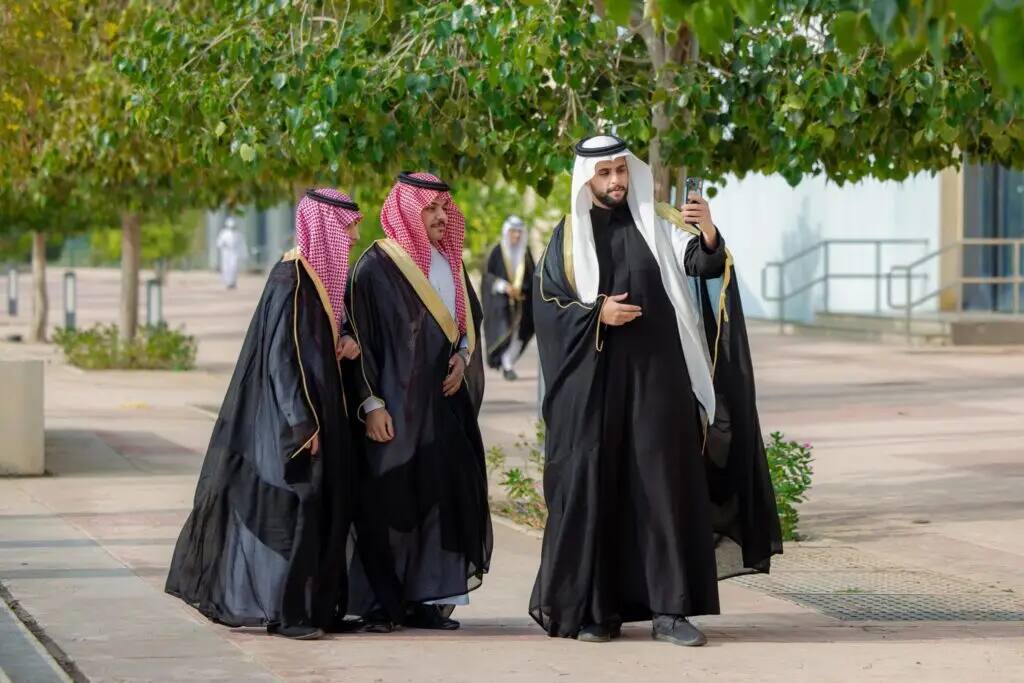 They are all three men in traditional Saudi dress, wearing the Saudi Shemagh or Ghutra, all graduates of Qassim University, and finally a selfie under the lush foliage on a sunny sidewalk. Two of them are smiling while the third is holding his cell phone to capture this special moment.