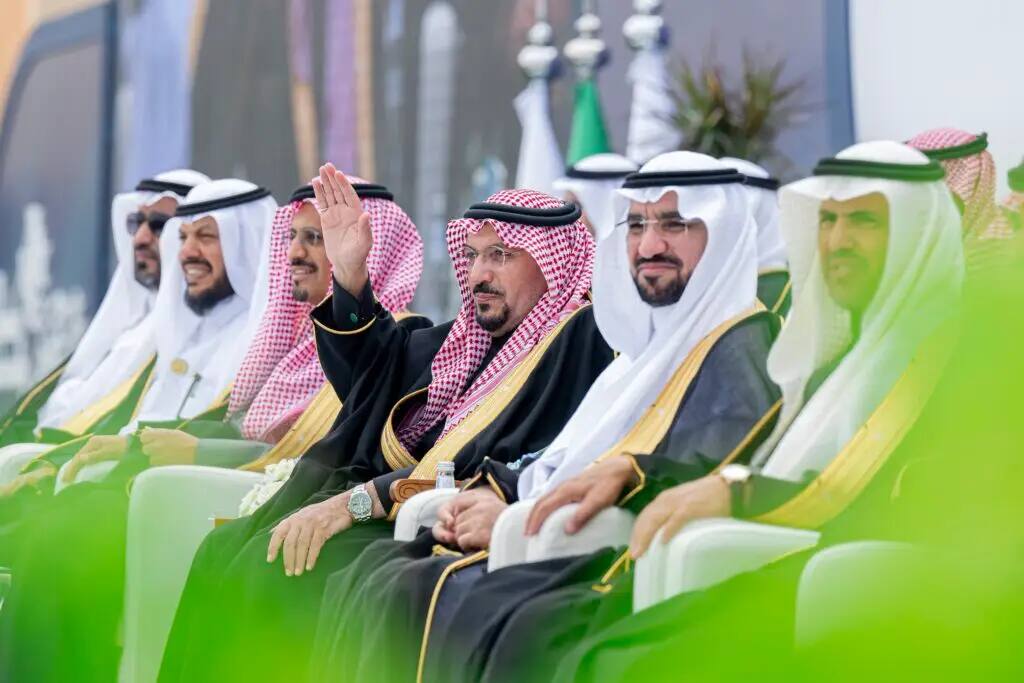 A group of men in traditional Saudi dress, including graduates from Qassim University, sit in the open air; he is described as greeting from the center while others watch attentively. Look for media and greenery in the background, giving the diverse material a safe and formal feel.
