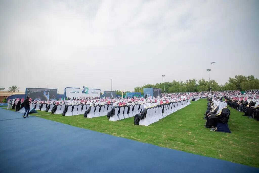 A large outdoor area gathers a large number of attendees, sitting in their books on a canvas background, sharing the traditional Saudi dress, draped in the thobe and shemagh or Saudi ghutra. The venue is adorned with banners and huge screens celebrating the university's graduates, and the honor is accompanied by His Highness the Emir of Qassim Province, until the cloudy weather sets in.