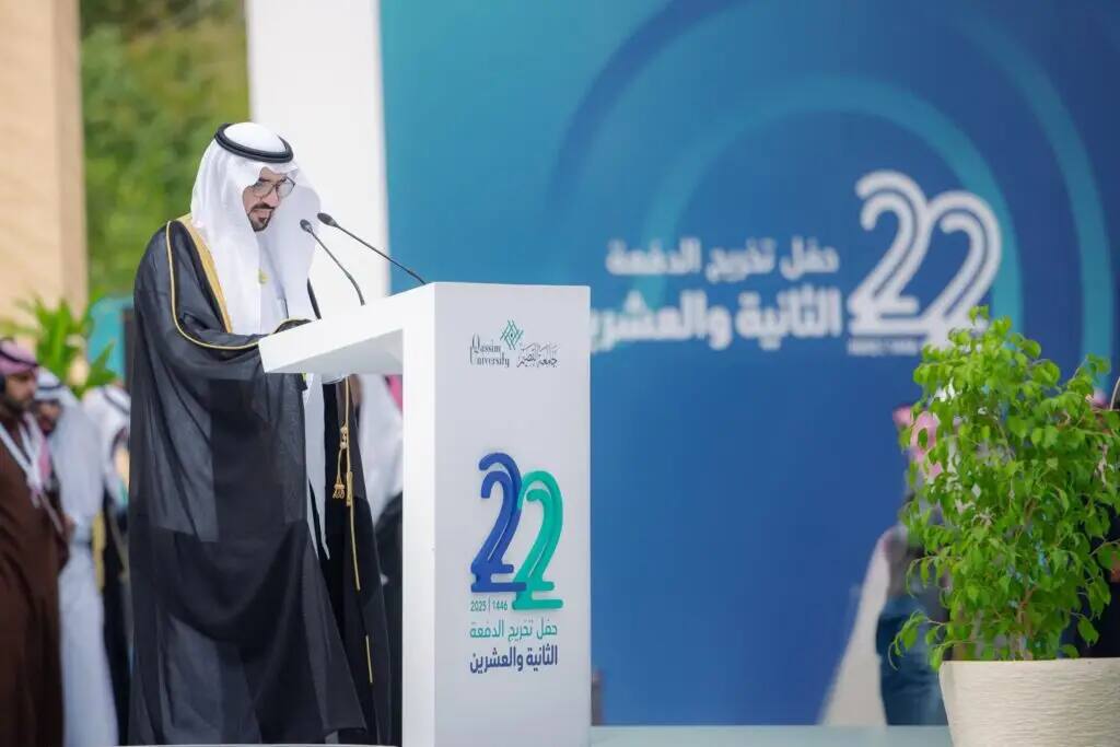 A man dressed in traditional Saudi attire with a Saudi shemagh at the speaking platform during the 22nd Qassim University Graduation Convocation, looking at a large blue board with the number 22 in both Arabic and English.