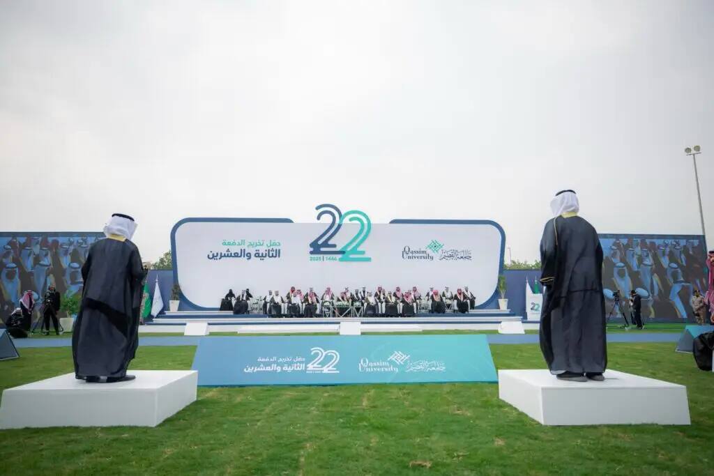 Held on a platform at Qassim University, decorated after "22", a number of dignitaries sit on the outdoor platform, their faces dressed in traditional attire. The patroness of the ceremony, dressed in the black color of the graduates above six, wears a Saudi shemagh in a Saudi ghutra, under an overcast sky.