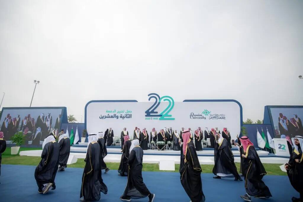 A group of people mention the official Saudi uniform, including the Saudi Shemagh or Ghutra, in front of a theater at an outdoor graduation. Large numbers (22) and Arabic text advise the age of the name "university" and honor "graduates" as part of the Qassim University events.