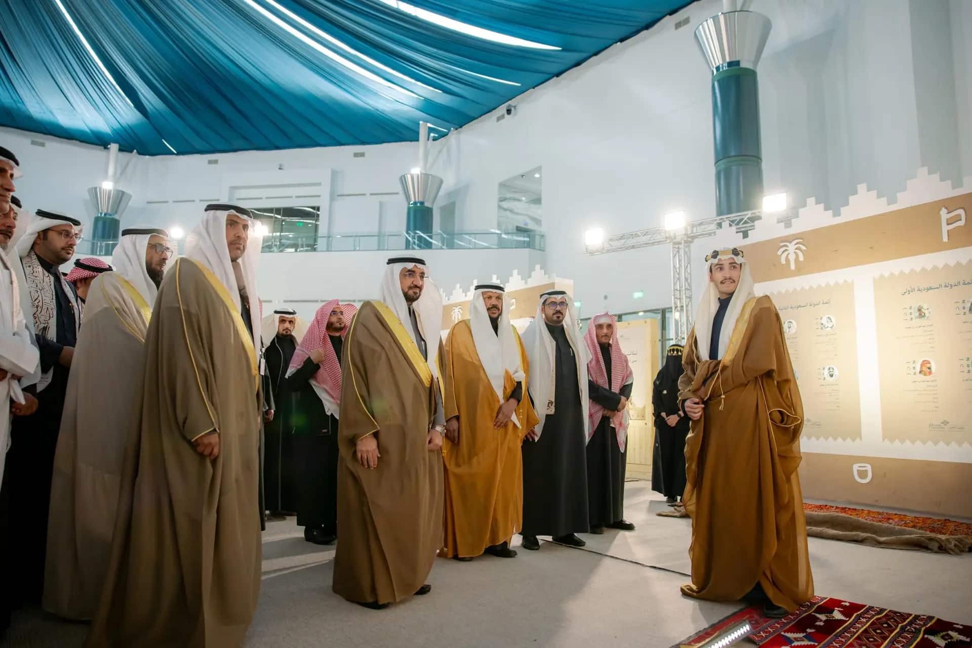 يجتمعون مجموعة من الرجال مرتدين الزي السعودي التقليدي، بما في ذلك معالي رئيس الجامعة، تحت سقف قناص بقماش الأزرق، حيث يتبادلون الأحاديث والابتسامات من خلال فعاليات يوم التنوع في مجتمع الرجال في القصيم.