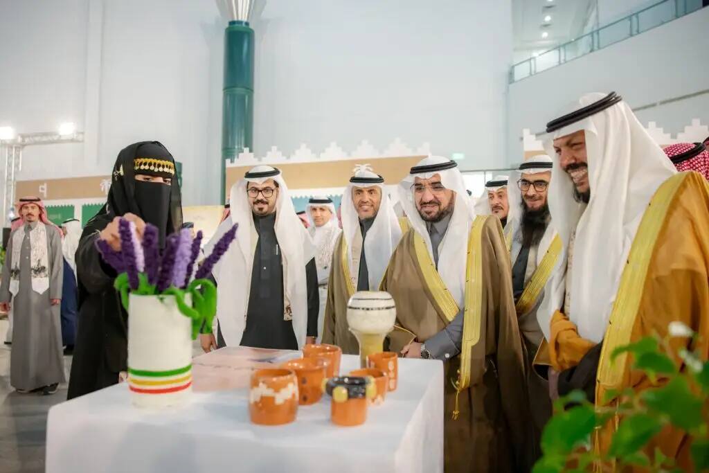 مجموعة من الرجال يخرجون الزي المباشر المباشر، من بينهم رئيس الجامعة، يبتسمون ويشاهدون سيدة ترتدي ملابس ونقاباً أسود وهي تقدم العاملين خزفية واناءً يحوي زهراً بنفسجية، وذلك من خلال فعاليات في إحدى القاعات ضمن معرض أو النقابة.