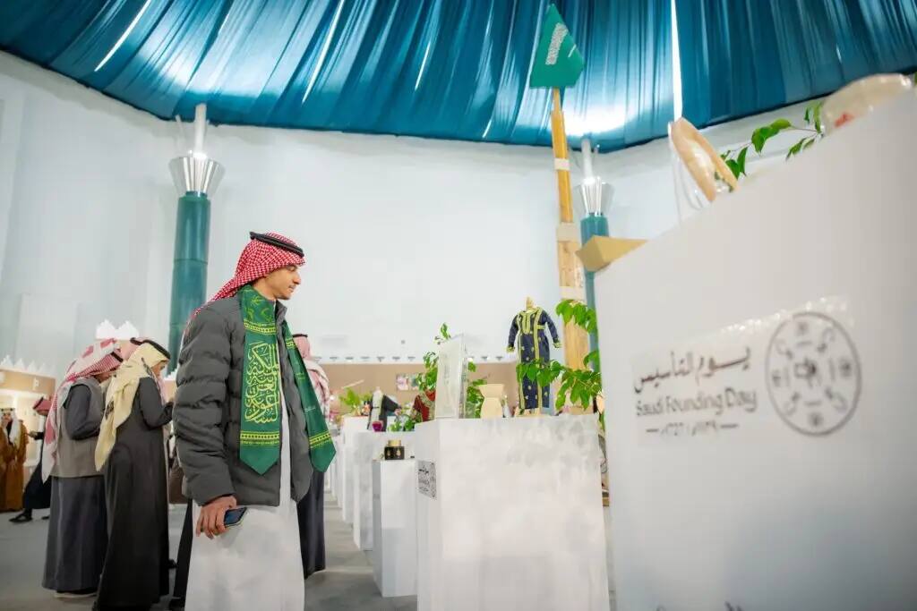 يقوم أحد الرجال مرتدياً الزي السعودي التقليدي الأخضر ومزيناً وثيقة، بتفقد الأجنحة داخل معرض يوم التأسيس المقامة الجامعة العريقة، حيث تظلل المكان مظلة زرقاء وتضفي الزينة الزينة من الثقافة السعودية وتعتبر مساهمةً على الحضور.
