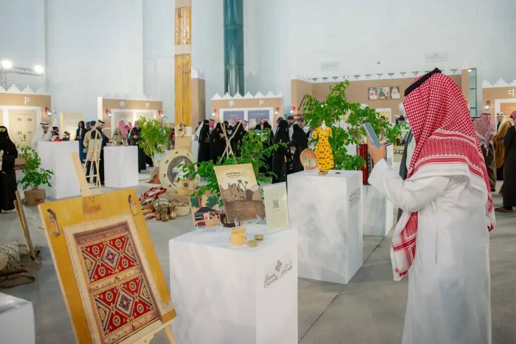 ارتدى رجل سعودي أصلي (الثوب مع الشماغ أو الغترة السعودية) يلتقط صورة بهاتفه المحمول خلال فعاليات يوم التأسيس، وذلك في معرض داخلي يضم معروضات من الحرف اليدوية الفنية والنباتات، وتميز في الخلفية لعدد من الزوار الآخرين.