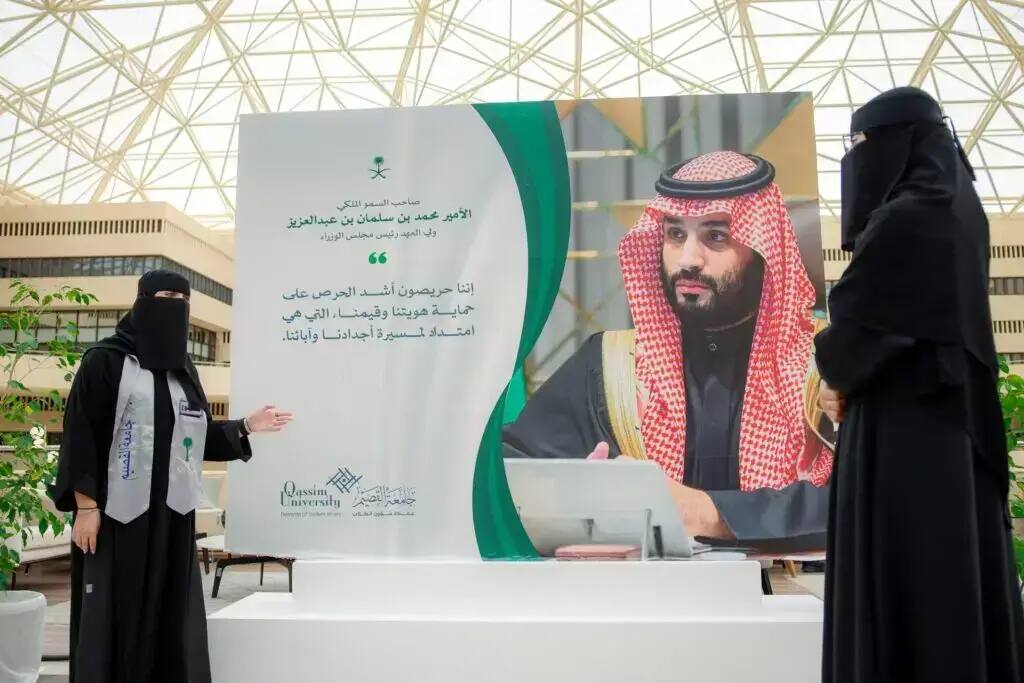 تتقف أمرأتان ترتديان ملابس بيضاء ونقاباً بجوار لوحة كبيرة لصاحب السمو الملكي الأمير محمد بن سلمان بن عبد العزيز آل سعود، تتضمن نصوصاً عربية وشعارات شعبية، وذلك في مساحة لا تتجاوز سقف زجاجي داخل الحرم الجامعي، مما يبرز مكانة العلم وروح يوم العلم في جامعة القصيم.