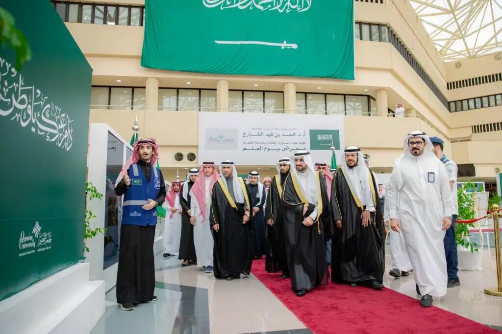 يسير عدد من الرجال، ويرتدي العديد منهم الشماغ أو الغترة السعودية التقليدية، على البساط الأحمر داخل مبنى كبير تتفوق عليه راية المملكة العربية السعودية في مشهد عكسي مكانة يوم العلم. شارك بعض المشاركين في بطاقة التعريف الرسمية، بينما خلفهم مجموعة من المتابعين والصورين في الخلفية.