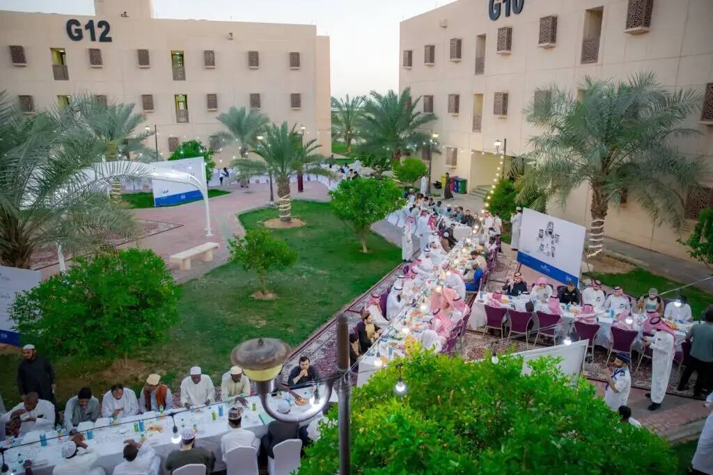 A large number of participants gathered in the Partnership Square with buildings numbered G10, G12, and palm trees, around long tables, sharing the month of Ramadan in a special Ramadan, marking the end of the Al Rayyan Ramadan program in Qassim. The event was characterized by a spirit of familiarity and community participation, with attendees dressed in Saudi national attire, including the Saudi Shemagh and Ghutra.