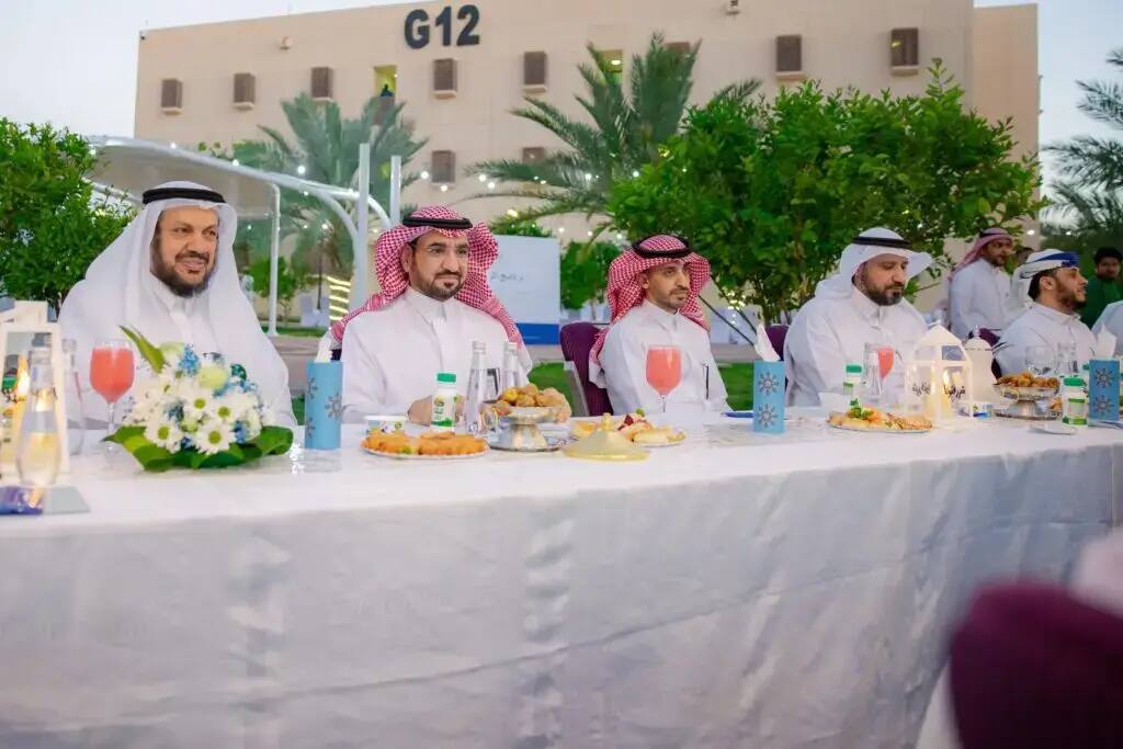 A number of men dressed in Saudi uniforms, including a shemagh or Saudi ghutra, led by the university president, gather around a long table with a white tablecloth in the open air. The table is decorated with a variety of foods and lanterns, as part of the Al Rayyan Ramadan program. To search the large green spaces and the G12 building of Qassim University.