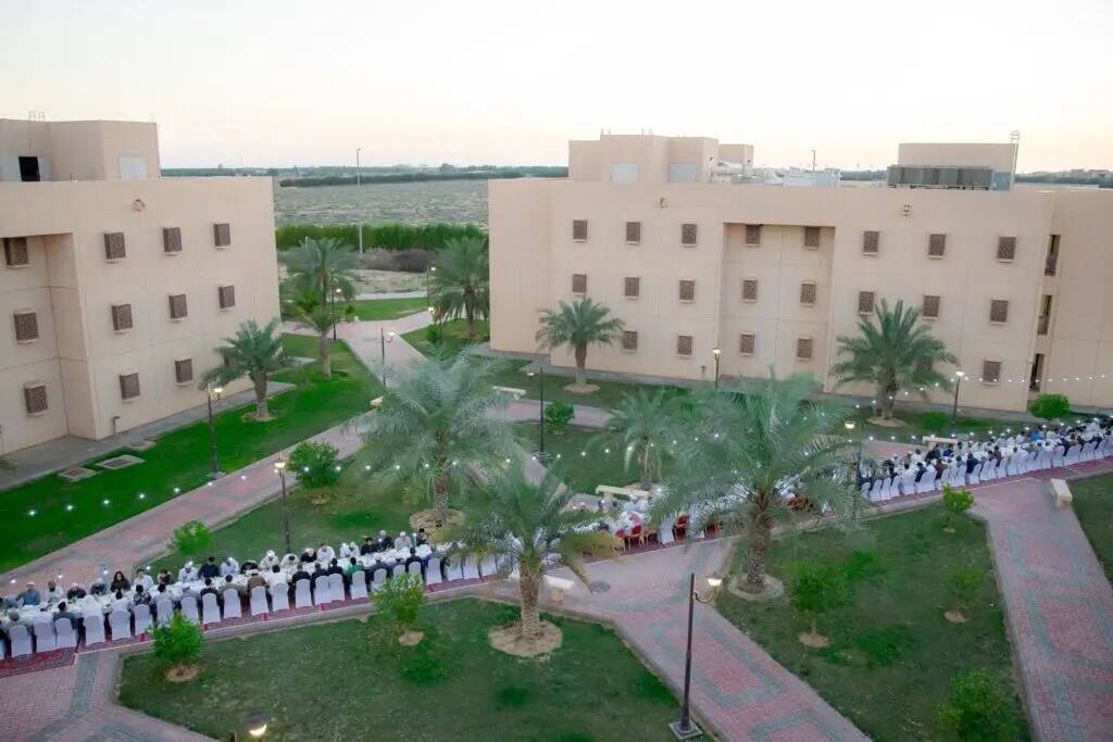 A large gathering of people, dressed in traditional white dress with the Saudi shemagh or ghutra, gather at a long outdoor table between commercial buildings and palm trees. The sunset Iftar gathering is the culmination of Qassim University's Al-Rayyan Ramadan program.