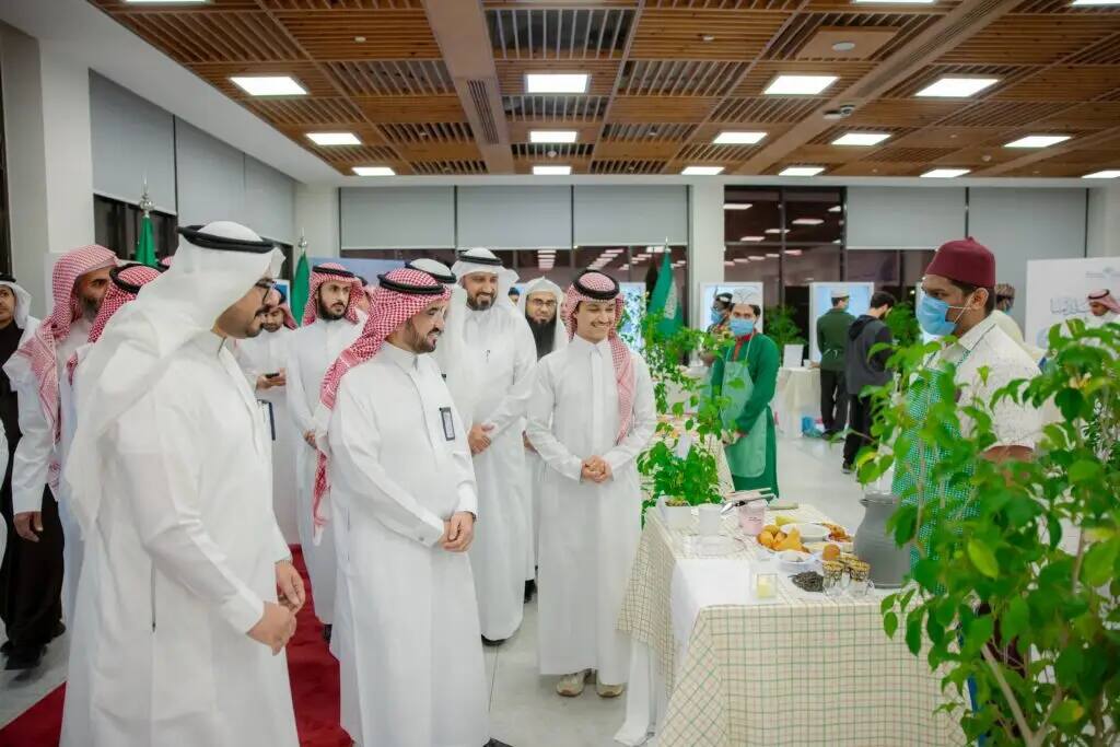 A group of men wearing the Saudi Shemagh or Saudi Ghutra gathered around a table with a full menu and food items, listening to the President of Qassim University wearing a mask, as he gave a presentation at an indoor event as part of the Al-Rayyan Ramadan program.