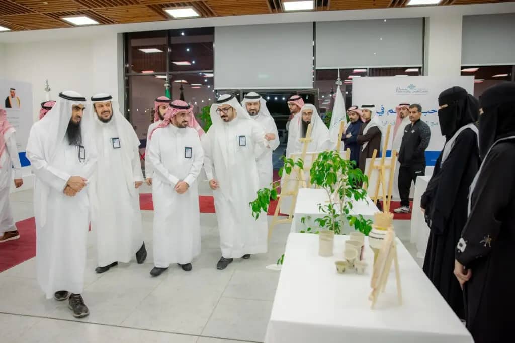 A group of men in the classic Saudi garb of thobe and shemagh or Saudi ghutra walk around an exhibition with floral paintings for display, during the Al-Rayyan Ramadan program. Another is the group of women in black robes. The event takes place in a brightly lit indoor hall as part of the official Qassim University activation.