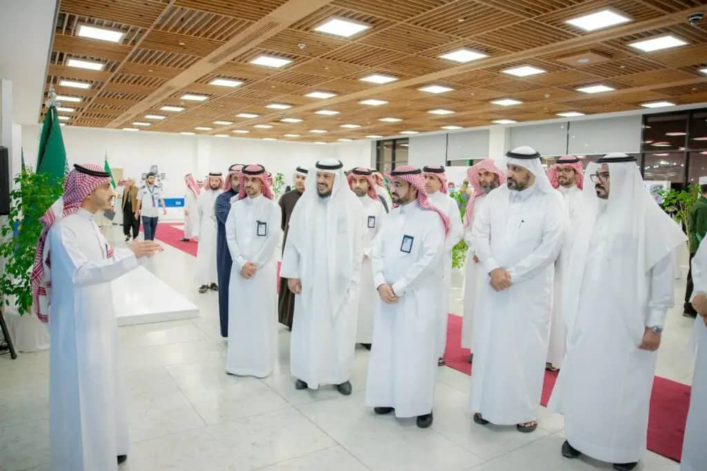 Inside a hall with a modern design and a wooden ceiling, men wearing the Saudi shemagh or tunic listen intently to the university president during the conclusion of the program. The venue is brightly lit, with plants and signs of Saudi Arabia, a perfect and symbolic representation of Qassim University.