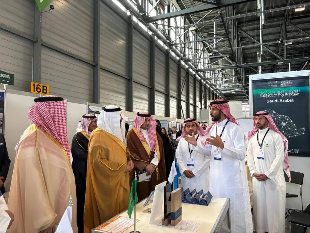 A number of men dressed in traditional Saudi attire, including a shemagh or Saudi ghutra, gather around the Qassim University pavilion at the Geneva International Exhibition of Inventions. One of the attendees is explaining and demonstrating, while enjoying the focus and attention. He displays airplane tires bearing the Saudi Arabia logo, as well as bronze stickers for its airlines.