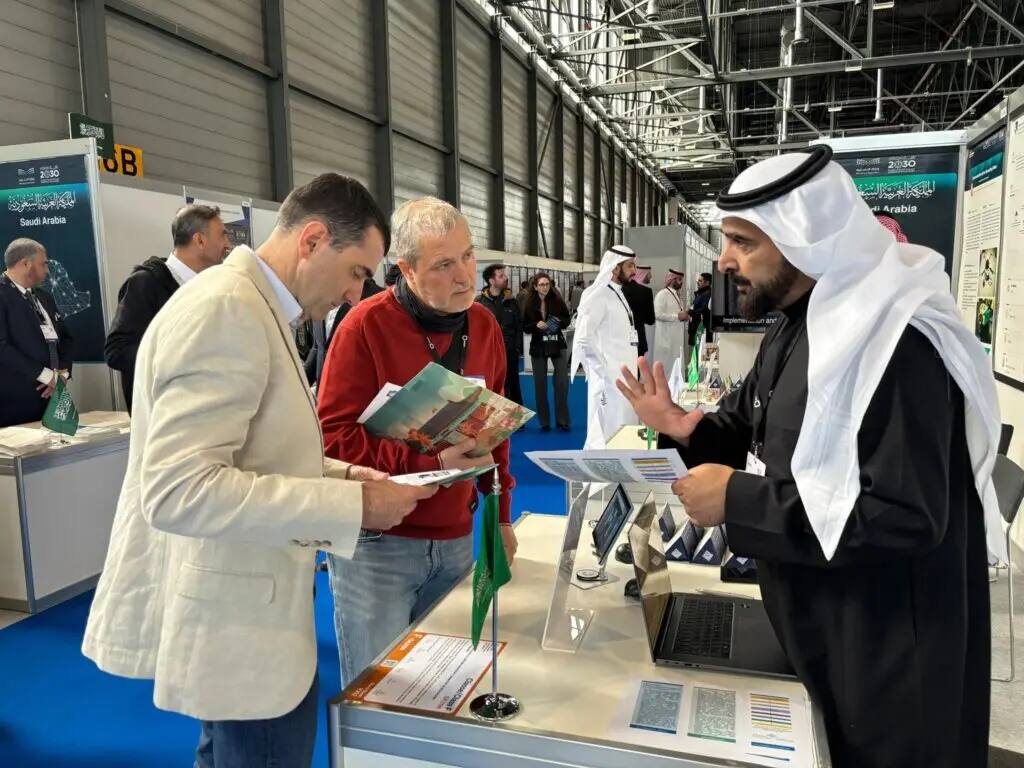 Three men interact at the Qassim Center's exhibition booth at the Geneva International Exhibition of Inventions. One man, dressed in a traditional Saudi mother's dress with a Saudi shemagh, is seen interacting with the two men while holding informational pamphlets. A number of visitors and other exhibition pavilions can be seen in the background.