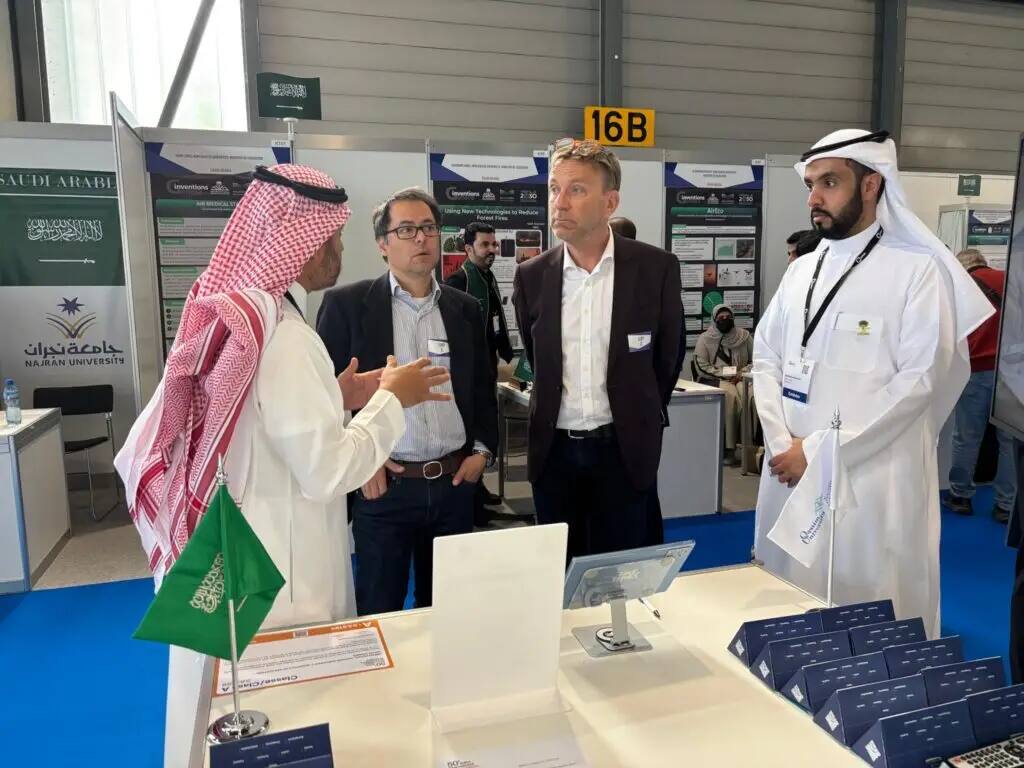Four men are conversing at the Qassim University pavilion, two of them in the Saudi uniform of shamagh and national dress, and two in suits. The pavilion contains brochures and Saudi Arabian flags, as well as presentations about the bronze medals the university won at the Geneva International Exhibition of Inventions.