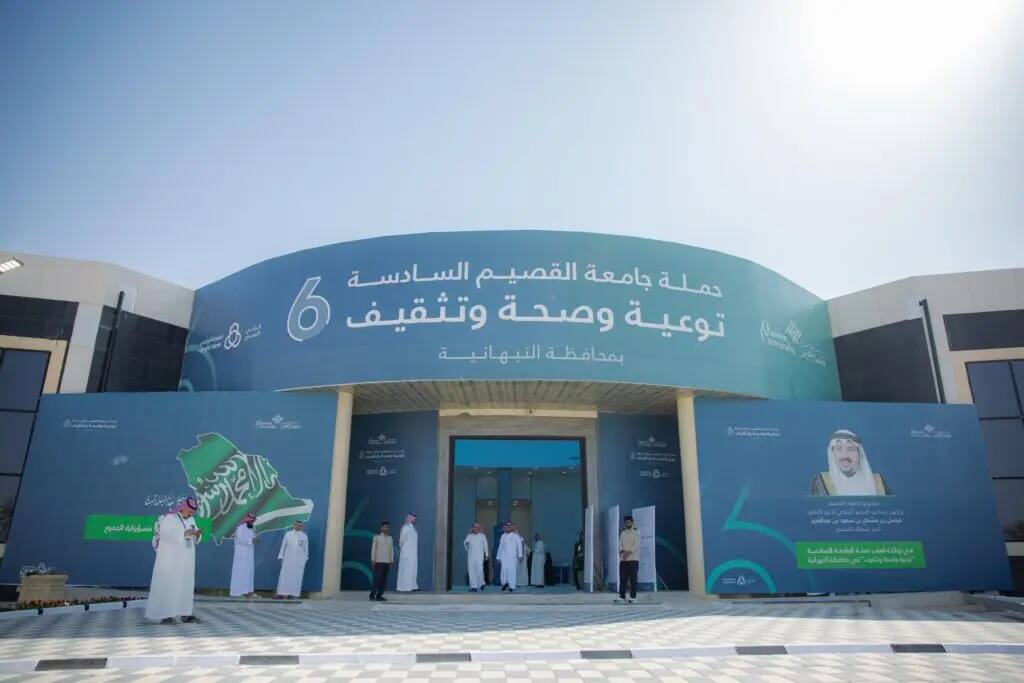 واجهة مبنى كبير أزرق يحمل نصوصاً وشعارات باللغة العربية، ويتواجد أمامها مجموعة من الأشخاص الزي السعودي الرسمي، من شماغ سعودي أو غترة سعودية مع عقال وبشت سعودي. وقفت لافتة تحمل علم المملكة وصورة الشخصيات المميزة، في إطار حملة توعوية وصحية تنظمها جامعة القصيم.