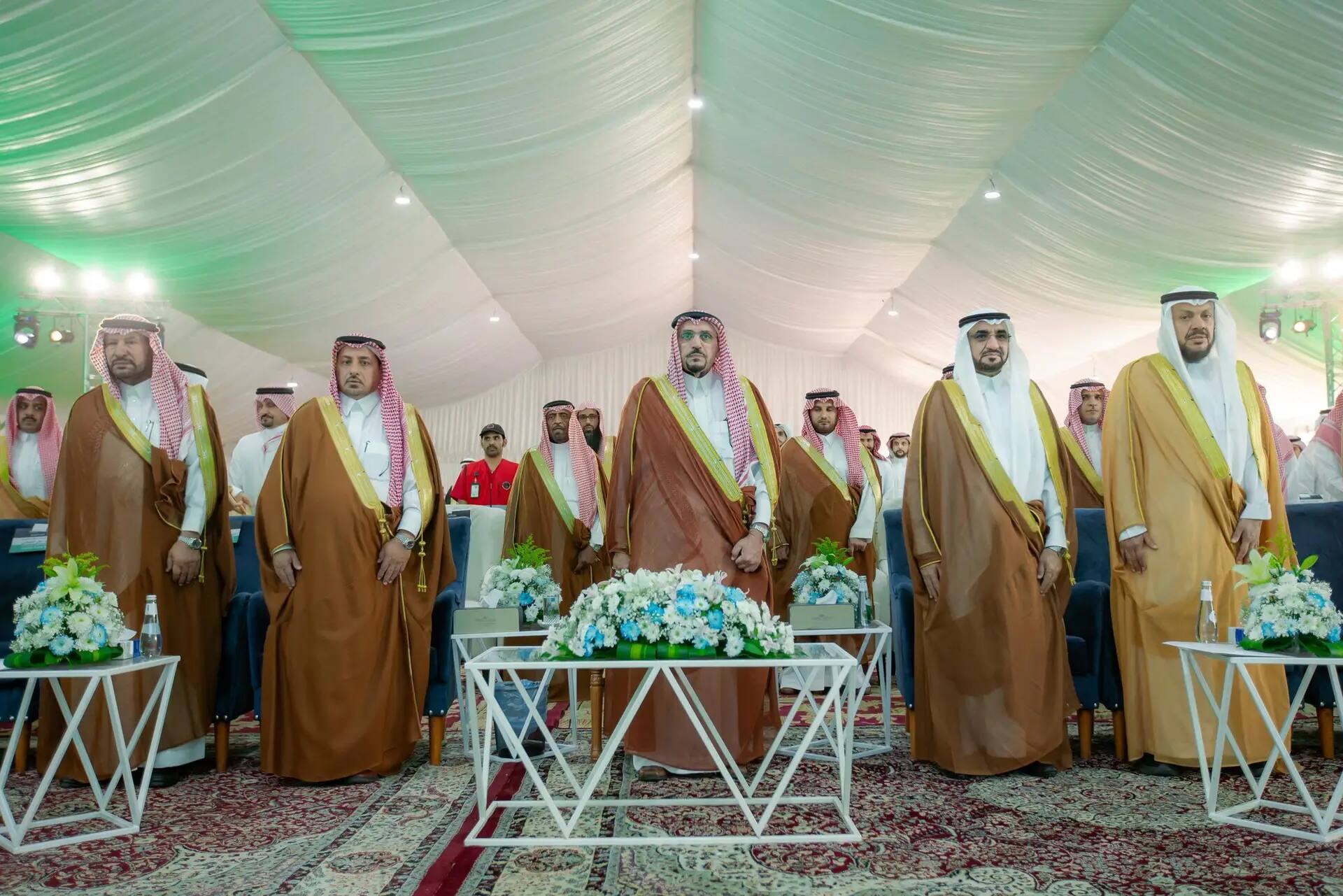 A group of men wearing the Ghutra or Shemagh and Eqal stood in front of the Saudi Bisht, a tent decorated with white curtains, flower arrangements and decorative carpets, during a ceremony attended by His Highness the Emir of Qassim Province as part of the comprehensive university campaign in Nabhaniyah Governorate.