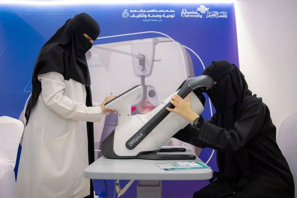 Two women, dressed in abaya and Saudi bisht with ghutra and headband, perform a medical eye exam with an expert at a clinic. In the background, a blue-colored awareness board with Arabic texts on the topic of health, as well as the Qassim University logo, highlights the healthcare efforts offered in Nabhaniya province.