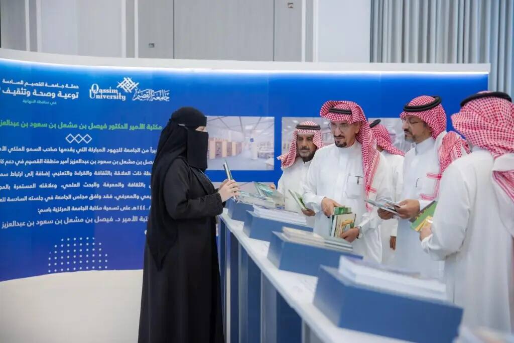A white lady and niqab, to a group of men in white robes, red Saudi shemaghs, and a Saudi aqalasht, at an informational corner in Qassim. This corner aims to promote awareness and education among visitors through blue banners and various books displayed in an organized manner.