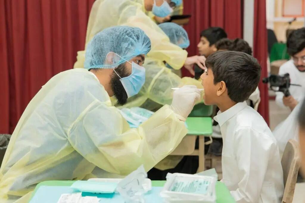 A health staff member, wearing his medical kit and mask, takes a swab from a child wearing a white shirt during a medical examination for pediatric health awareness, as part of a children's event in Qassim. Note in the background a number of indigenous staff, and the participating staff are wearing the Saudi Ghutrah and Eqal or Saudi Shemagh with the Saudi Basht in line with the university's nationality and official appearance.