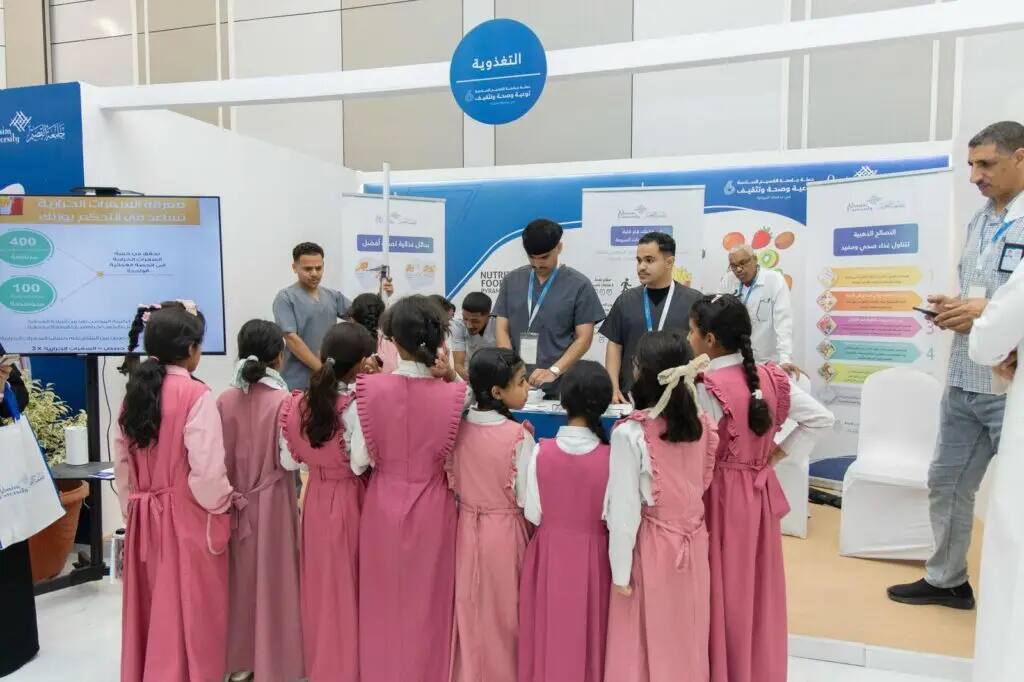 A Miami group of female students, dressed in matching uniforms, in front of an informational pavilion featuring awareness posters and an interactive display screen. The students enjoy the attention of the presenters, who are dressed in the Saudi Bisht and Ghitra or Shemagh and Egal, as they provide educational lectures on public health and nutritional awareness as part of the Health and Nutrition Exhibition organized by Qassim University.