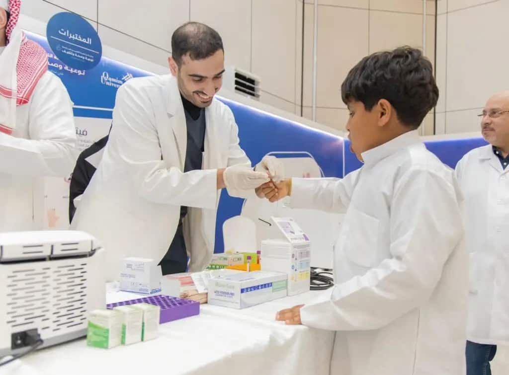 A healthcare worker, wearing a white coat and medical gloves, pulls a small child's dolls at the Health and Awareness Hospital in Nabhaniyah, as part of an event organized by Qassim University. Several medical supplies and boxes can be seen on the table. Some employees can be seen in the background, some of whom are wearing the Saudi shemagh and headband or the Saudi ghutra with the official bisht, reflecting the patriotism of the participants and the professional organization of the event.