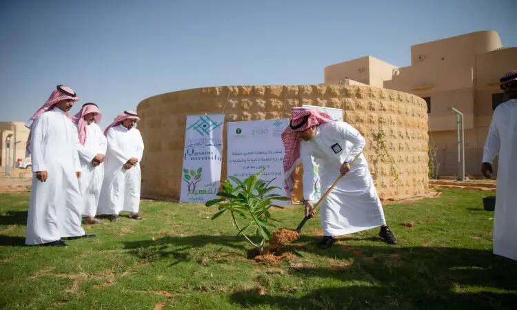 A small group of men take on the Saudi Arabian tunic or shemagh, aqal and besht, while someone plants a sapling in a tea area near circular buildings and banners bearing Qassim University's slogans about environmentalism and green initiatives.