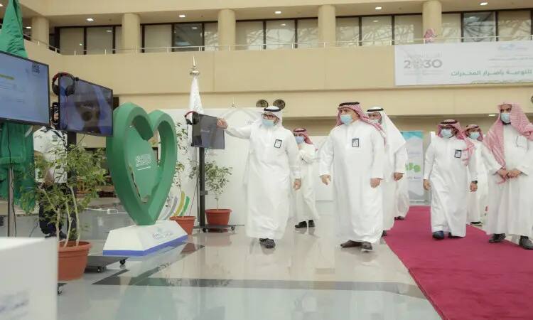 مجموعة من الرجال الثياب البيضاء، ويضعون الغترة أو الشماغ السعودي مع العقال، وبعضهم يرتدون البشت السعودي، يسيرون في الداخل بالقرب من زينة خضراء على شكل قلوب ونباتات مزروعة في أصص وشاشة عرض كبيرة، وذلك ضمن فعاليات معرض "واعي" للتوعية بأضرار المخدرات في جامعة القصيم. يُلاحظ أن بعض الحضور للكمامات، كما تلاحظون وسجادة ملونة ذات عنابي في الخلفية.