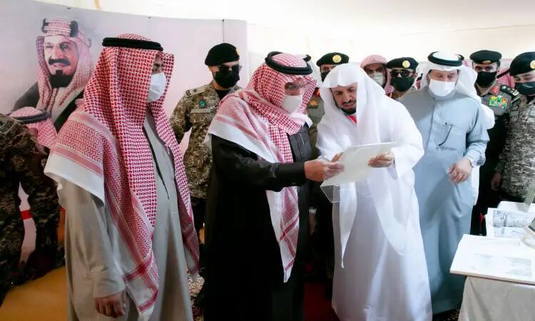 A group of men inside the university's pavilion, wearing the Saudi ghutra or shemagh, aqal and bisht, along with members in military uniforms, call out to each other. He is signing the attendance document, while in the background are large photos of Saudi Arabia's leaders and His Highness the Emir of Qassim Province.