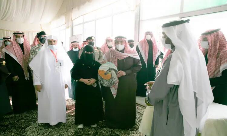 A group of people dressed in traditional Saudi attire inside the university pavilion during the Buraidah Spring Festival, where men are shown wearing the Saudi thobe and bisht and wearing the shemagh or ghutra and headband, while women are wearing abayas. One of the distinctive decorative pieces, and we notice the commitment of most of the attendees to wear masks in order to follow the approved health advice.